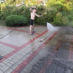 Fornalpol - Mężczyzna w krótkich spodenkach i klapkach spryskuje wodą z węża ciśnieniowego kostkę brukową z czerwonymi i szarymi elementami na tle zieleni.