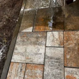 Mokre, wielokolorowe kostki brukowe ułożone w geometryczny wzór, z widocznym odpływem rynnowym w prawym górnym rogu i błotnistym brzegiem po lewej stronie.