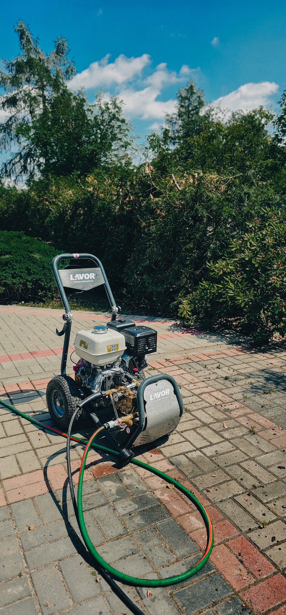 Profesjonalna myjka ciśnieniowa Lavor na kostce brukowej, z widocznym wężem i zielenią w tle, pod błękitnym niebem z chmurami.