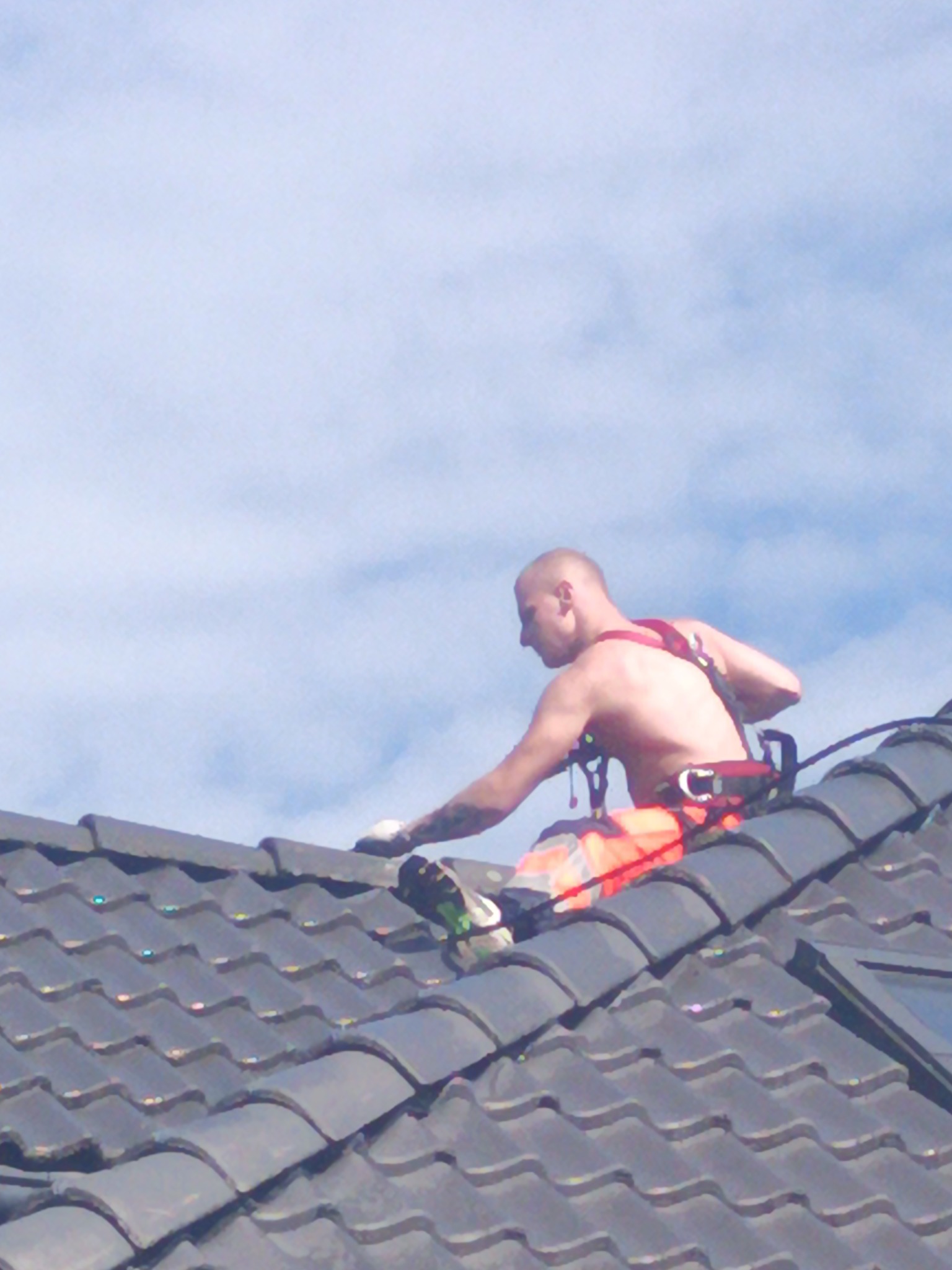 Pracownik bez koszulki w uprzęży zabezpieczającej, wykonuje prace na ciemnym dachu z dachówki ceramicznej, widok z dołu na tle jasnego nieba.