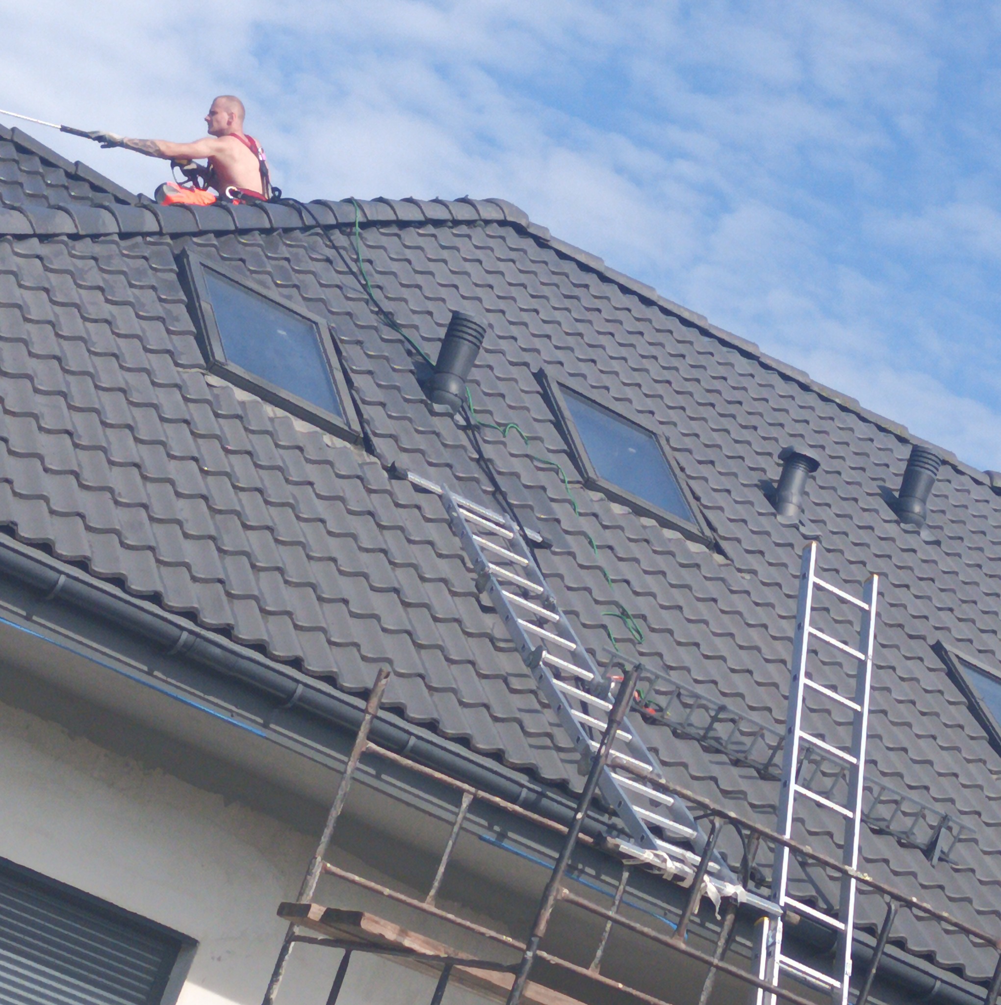 Pracownik w uprzęży, z długą lancą teleskopową, siedzi na ciemnoszarym dachu z oknami połaciowymi i kominami, na tle błękitnego nieba. Dwie drabiny oparte o dach.