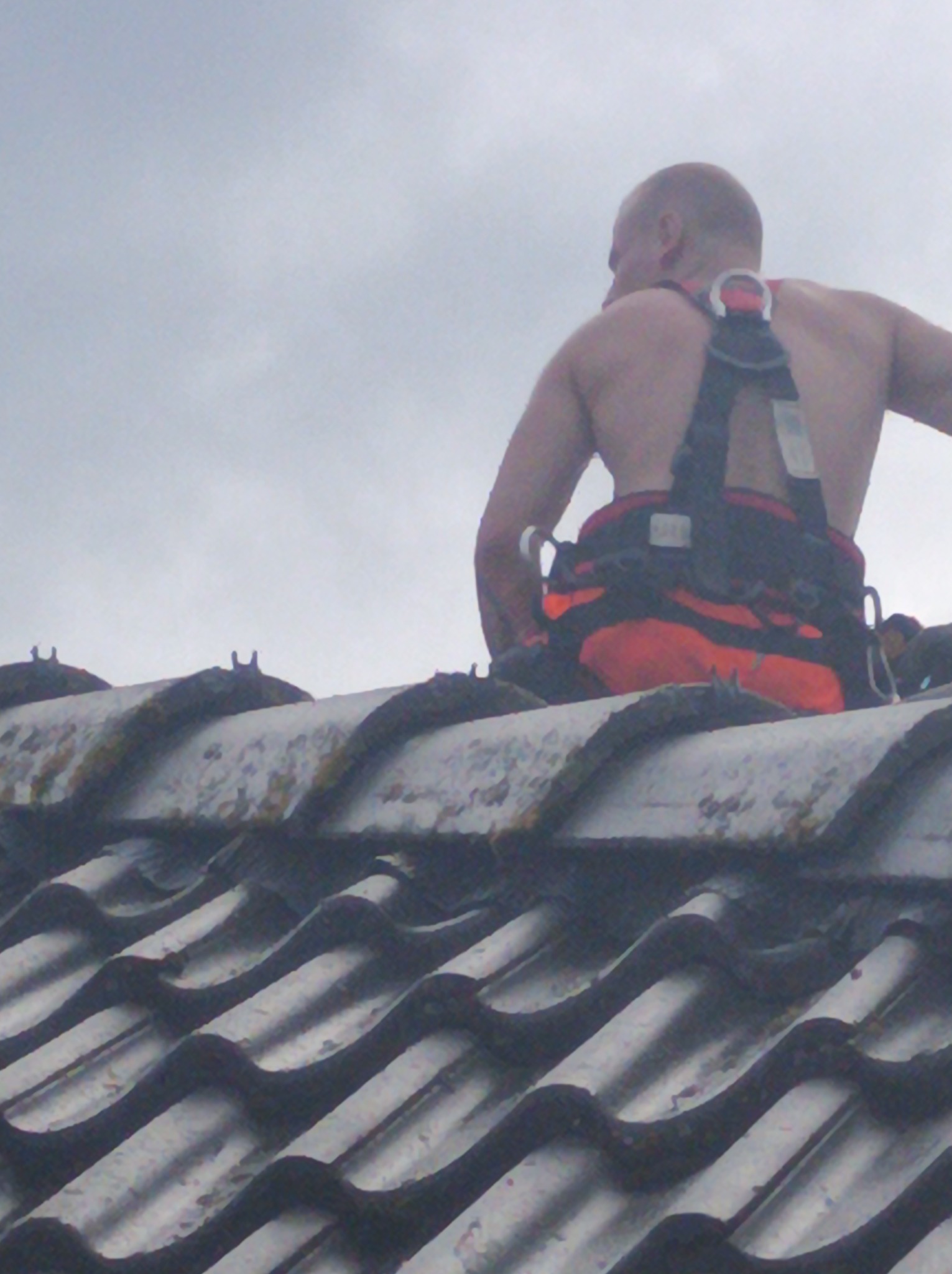 Pracownik w uprzęży, widok z tyłu, na spadzistym dachu pokrytym brudną dachówką w Tomaszowie Mazowieckim, pochmurne niebo w tle.