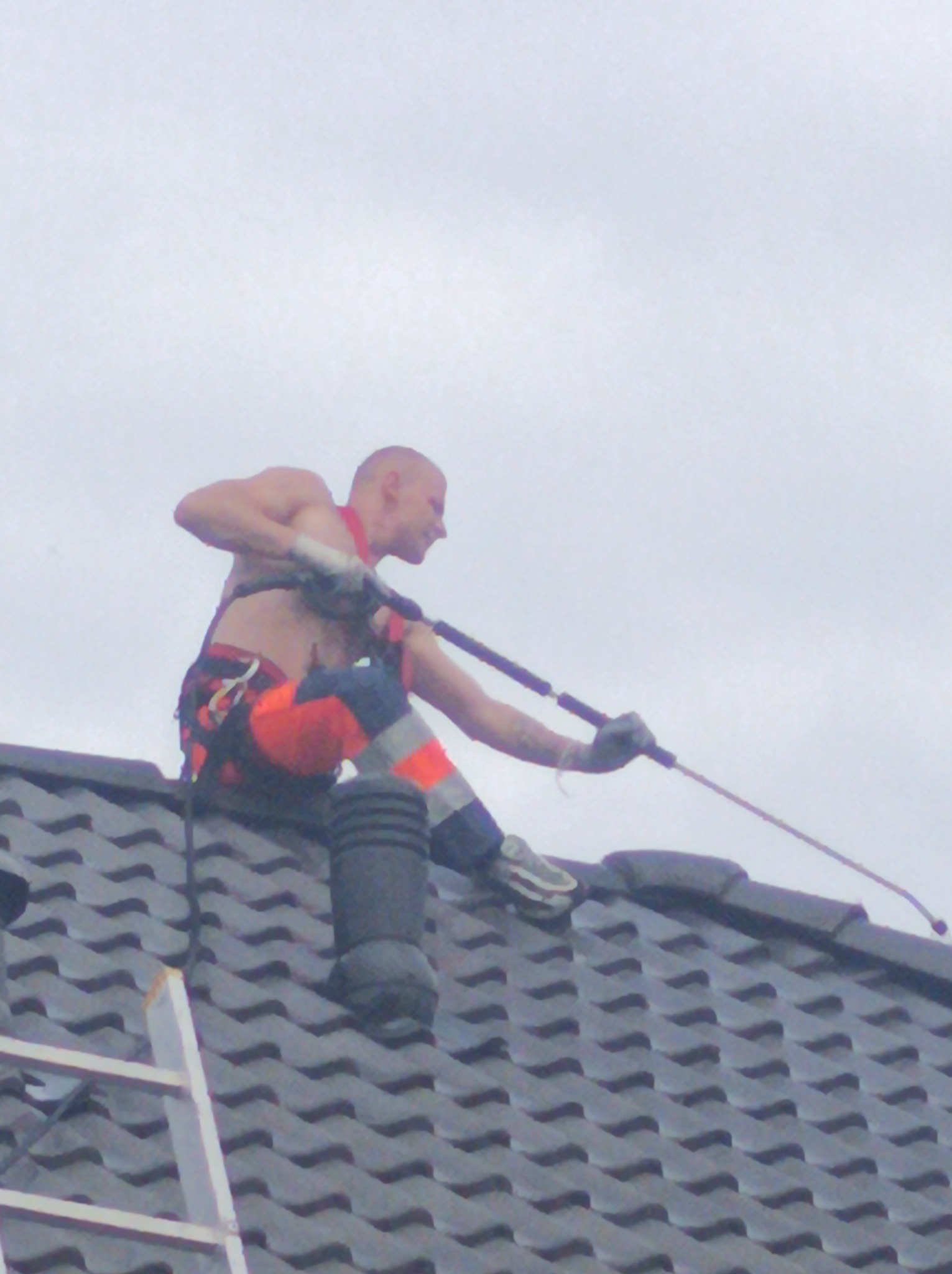Mężczyzna bez koszulki w uprzęży, z tatuażem na ramieniu, czyści dach z szarej dachówki za pomocą lancy ciśnieniowej; widoczna drabina oparta o dach.