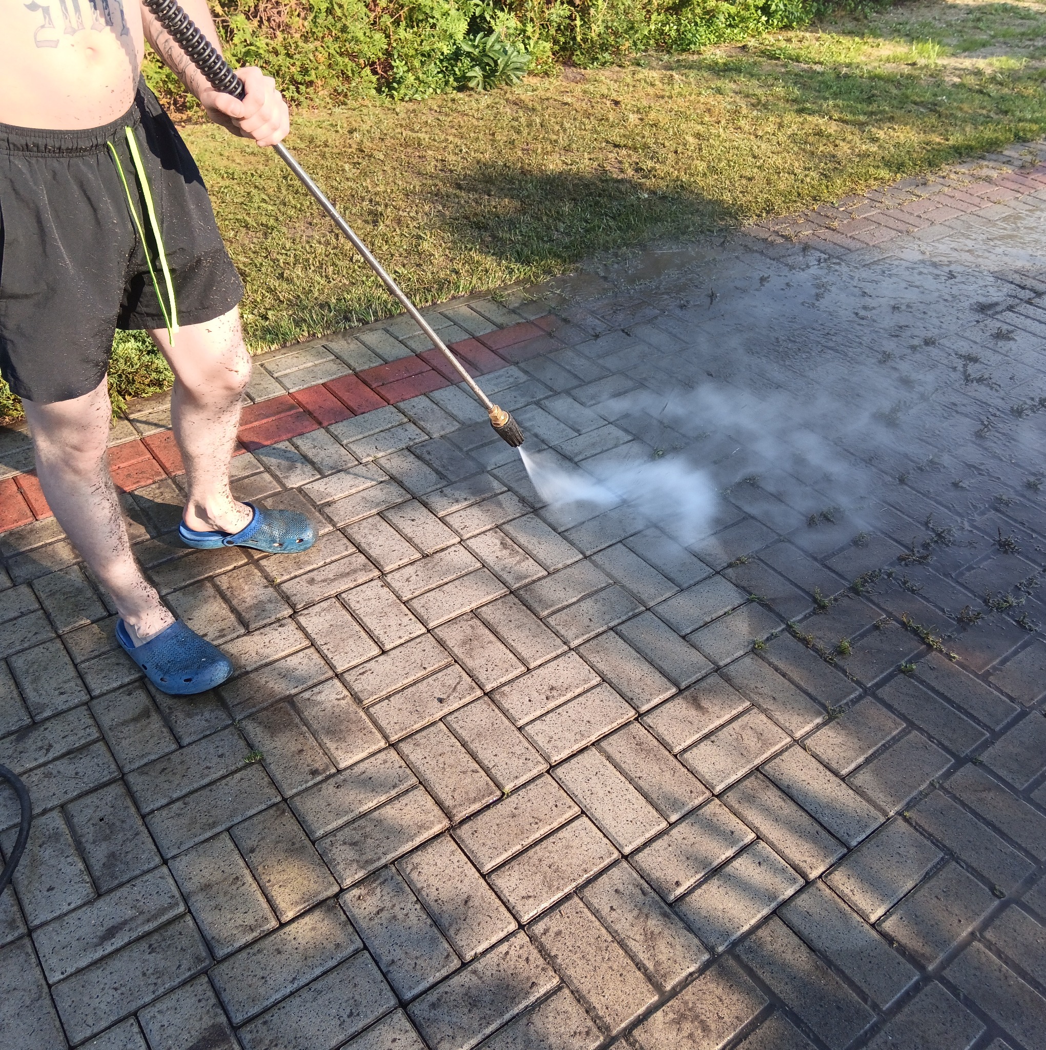 Mężczyzna w krótkich spodenkach i niebieskich chodakach czyści kostkę brukową za pomocą myjki ciśnieniowej. Widoczne zabrudzenia na nogach i kostce.