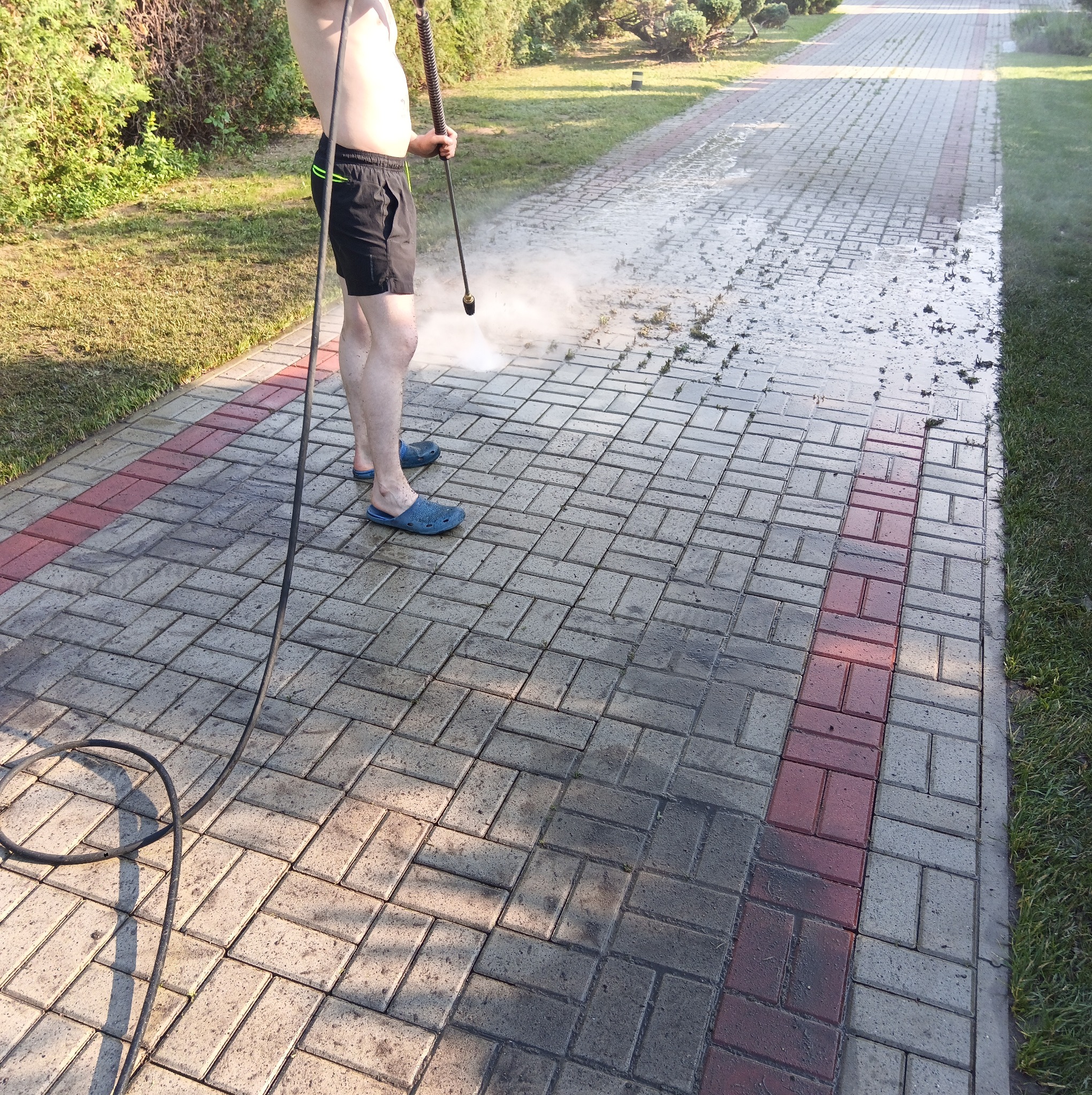Mężczyzna w klapkach i czarnych spodenkach czyści kostkę brukową myjką ciśnieniową na zewnątrz, widoczny efekt czyszczenia.
