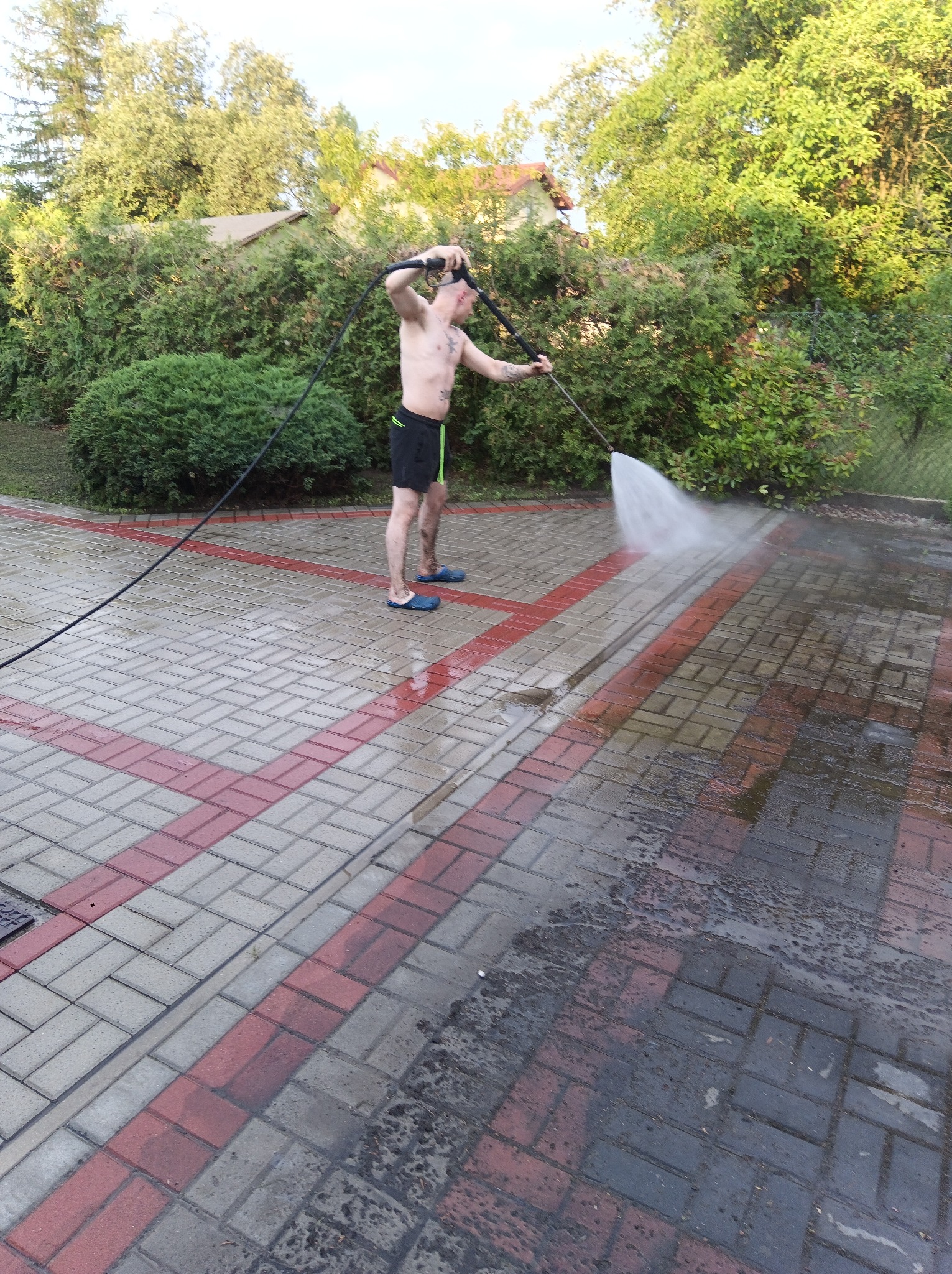 Mężczyzna w krótkich spodenkach i klapkach spryskuje wodą z węża ciśnieniowego kostkę brukową z czerwonymi i szarymi elementami na tle zieleni.
