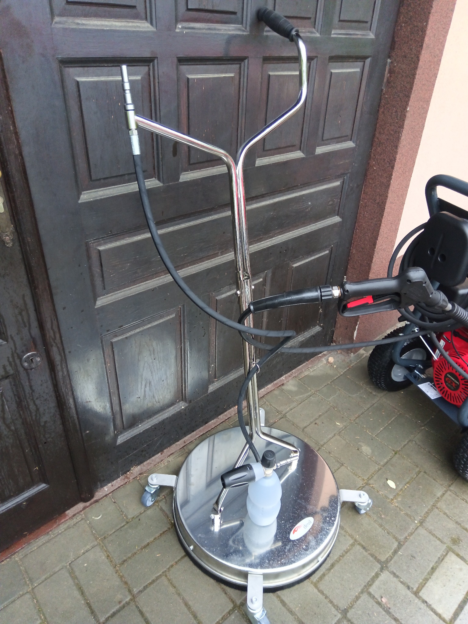 Profesjonalna myjka ciśnieniowa z okrągłą dyszą powierzchniową na kółkach, z podłączonym zbiornikiem na detergent, stojąca na kostce brukowej przed ciemnymi drzwiami wejściowymi i czerwoną myjką...