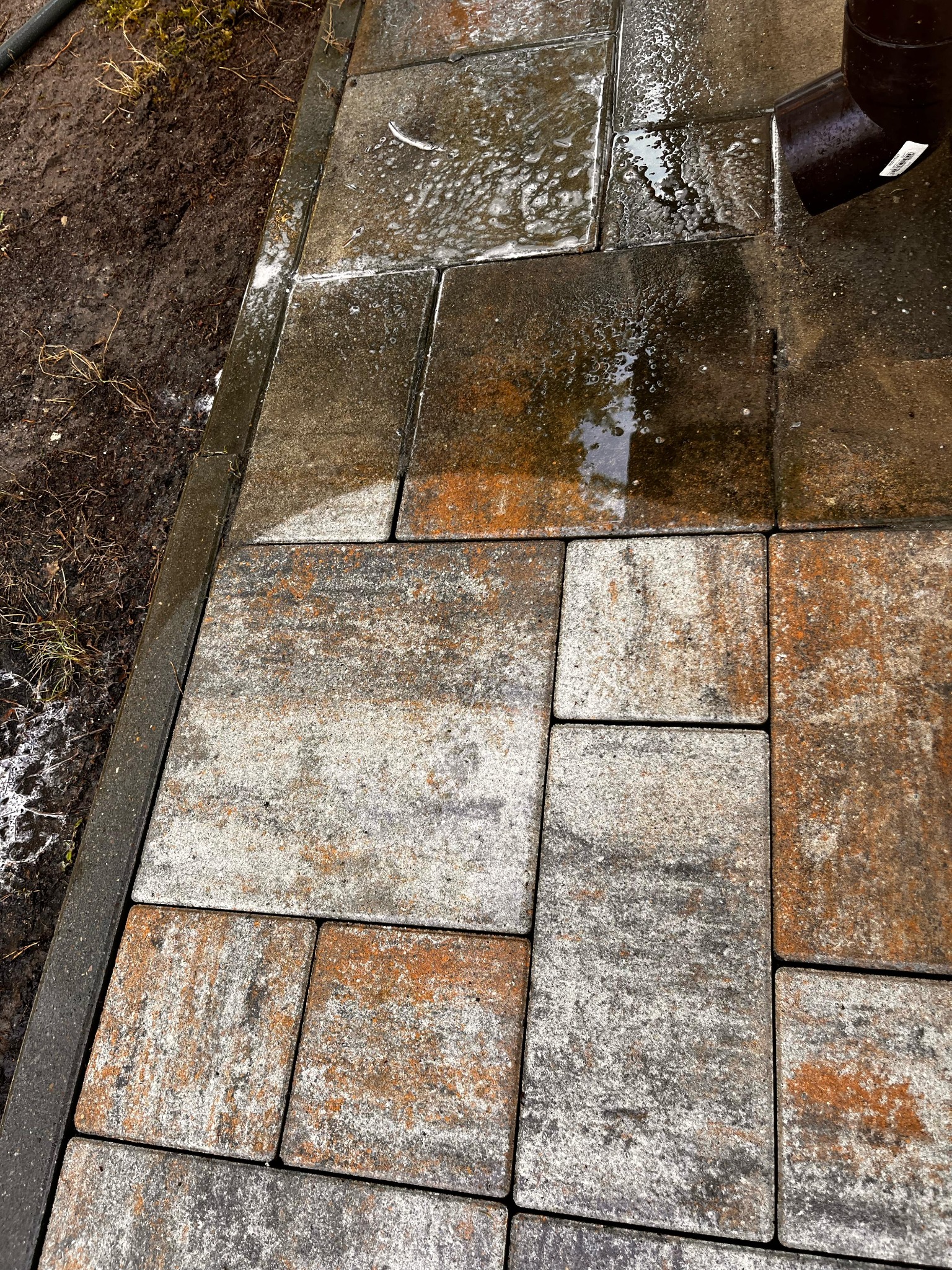 Mokre, wielokolorowe kostki brukowe ułożone w geometryczny wzór, z widocznym odpływem rynnowym w prawym górnym rogu i błotnistym brzegiem po lewej stronie.