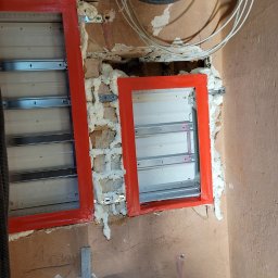 Elektromonter - Montaż dwóch czerwonych ramek pod rozdzielnice elektryczne w surowym stanie, widoczne przewody i pianka montażowa.