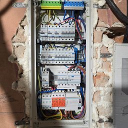 Elektromonter - Otwarta skrzynka rozdzielcza z zainstalowanymi bezpiecznikami i przewodami, zamontowana w surowej ścianie z widoczną cegłą i pianką montażową.