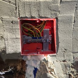 Elektromonter - Otwarta, czerwona puszka elektryczna z podłączonymi przewodami, umieszczona w surowej ścianie z widocznym tynkiem i pianką montażową.