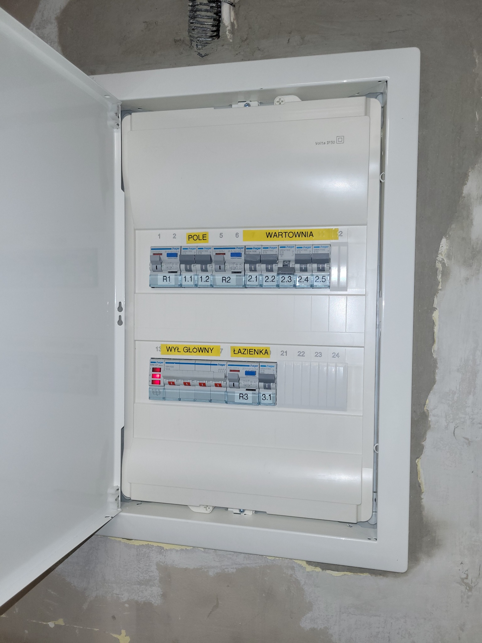 Otwarta skrzynka elektryczna Volta IP30 z bezpiecznikami Hager, widoczne oznaczenia 'Pole', 'Wartownia', 'Wył. Główny', 'Łazienka', zamontowana na surowej, nieotynkowanej ścianie.