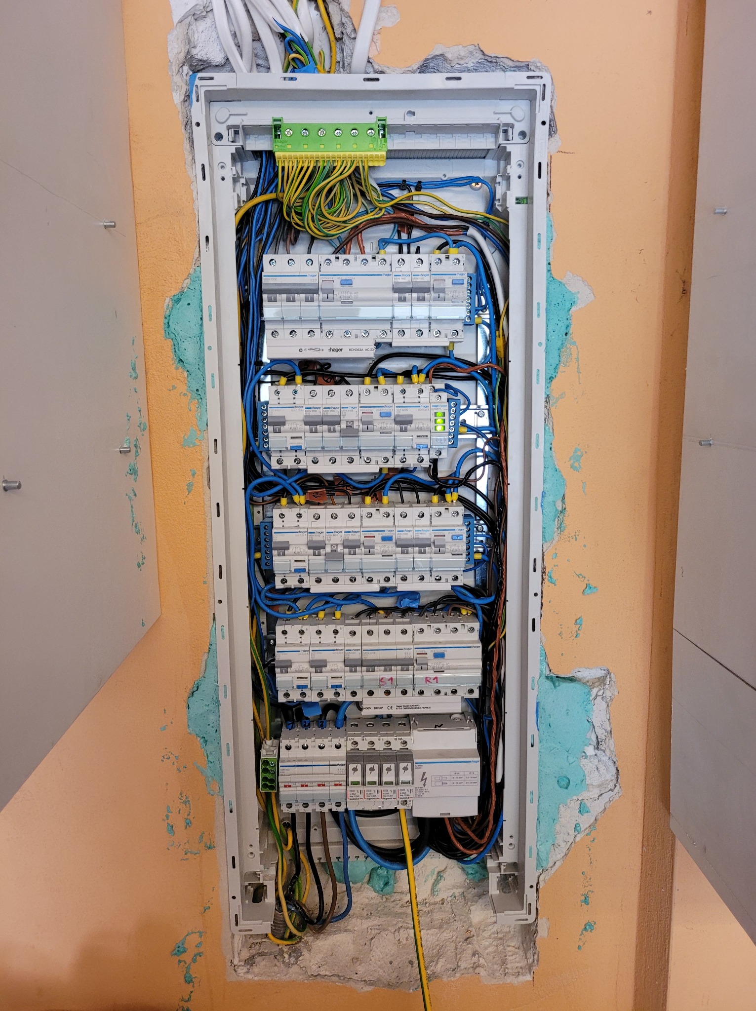 Rozdzielnica elektryczna Hager zamontowana w ścianie z widocznymi śladami pianki montażowej i przewodami w kolorach niebieskim, żółtym i zielonym.