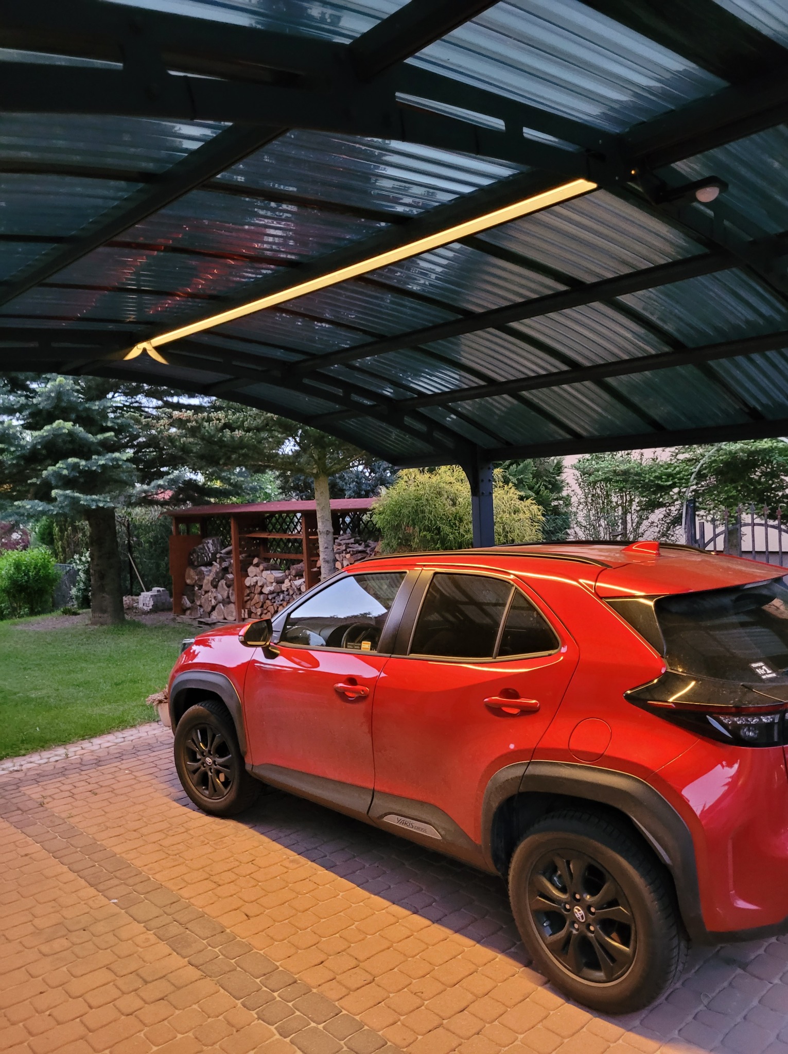 Czerwony samochód zaparkowany pod zadaszeniem z oświetleniem LED, widok z boku na kostce brukowej.