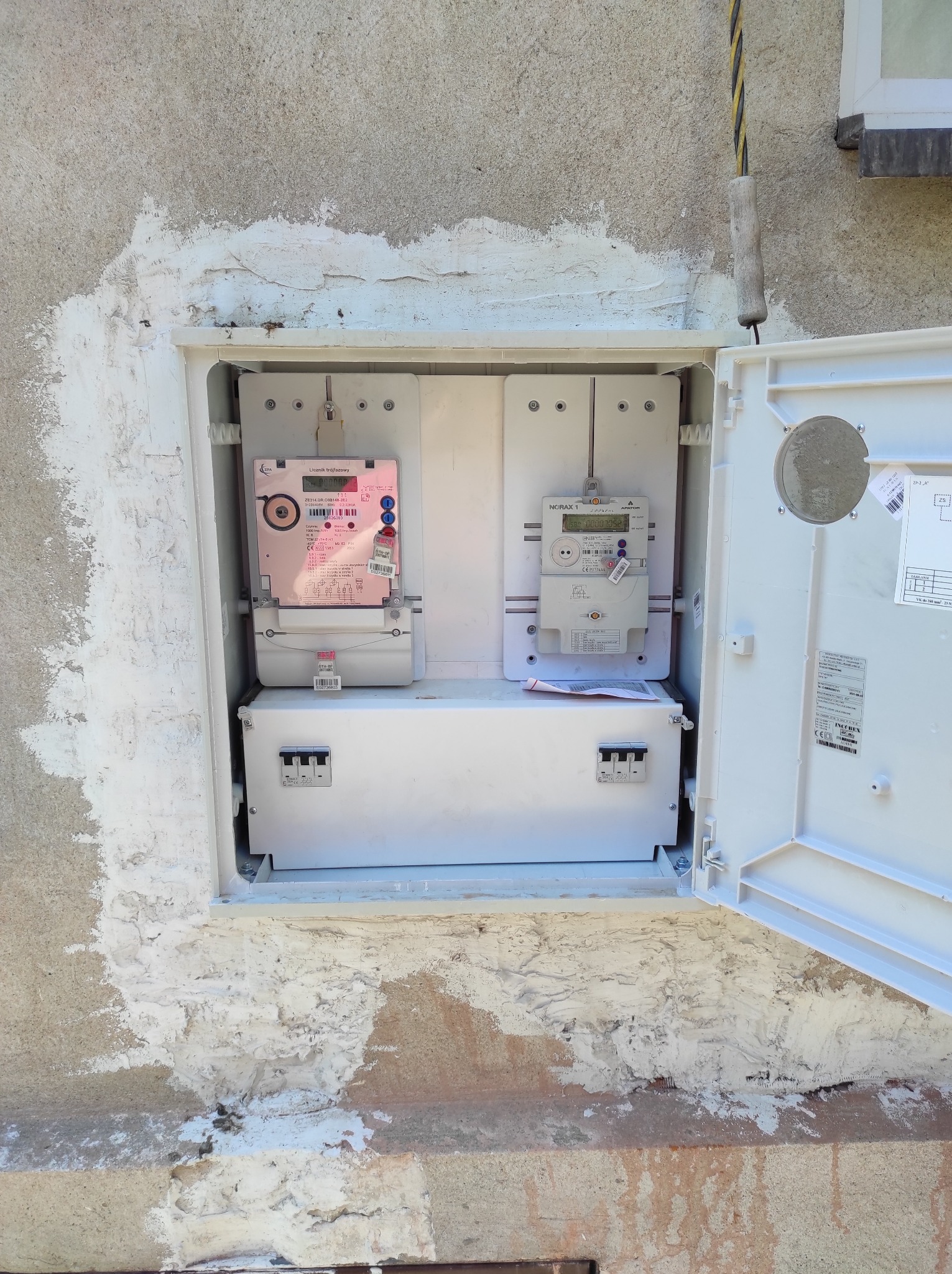 Otwarta skrzynka elektryczna z dwoma licznikami i zabezpieczeniami w Krakowie, osadzona w ścianie z widocznymi śladami niedawnego montażu i uszczelniania.