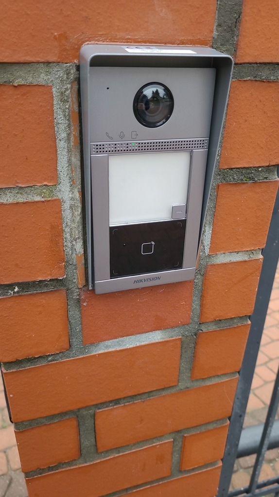 Szary wideodomofon Hikvision zamontowany na ceglanym słupku ogrodzenia, widoczna kamera, panel dotykowy i czytnik kart zbliżeniowych.