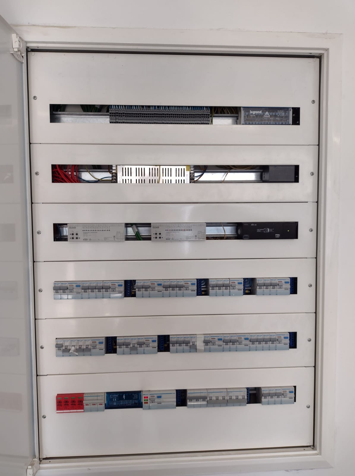 Biała skrzynka rozdzielcza z widocznymi bezpiecznikami, przewodami i innymi elementami instalacji elektrycznej, zamontowana w białej ścianie.