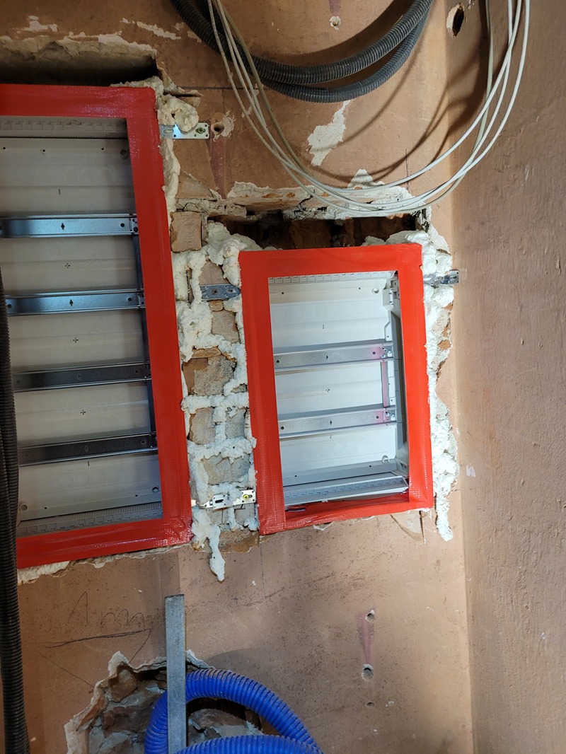 Montaż dwóch czerwonych ramek pod rozdzielnice elektryczne w surowym stanie, widoczne przewody i pianka montażowa.