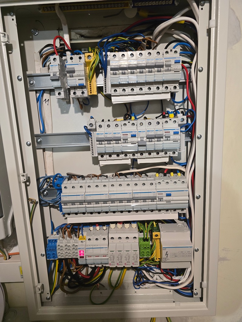 Otwarta skrzynka rozdzielcza z widocznymi bezpiecznikami, przewodami różnego koloru i elementami montażowymi na szynach DIN.