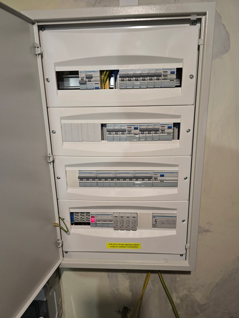 Otwarta skrzynka rozdzielcza z widocznymi bezpiecznikami, wyłącznikami i przewodami, zamontowana na surowej ścianie z widocznymi śladami po remoncie.