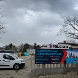 Zdjęcie przedstawia siedzibę hurtowni BHP i usług budowlanych Polgrid, z widocznym banerem reklamowym, białym samochodem dostawczym z logo firmy oraz budynkiem w tle.