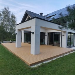 Paulina Witkowska - Nowoczesny taras kompozytowy z zadaszeniem przy domu jednorodzinnym. Jasne deski tarasowe, białe filary, duże okna przesuwne. Widok z poziomu trawnika.
