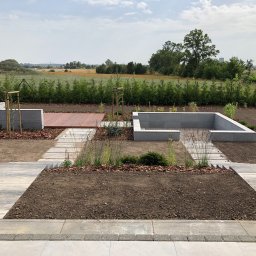 Karolina Dmitruk - Nowoczesny ogród w trakcie realizacji, widoczne podwyższone rabaty z szarego betonu, drewniany podest, ścieżka z płyt betonowych, młode drzewka podparte bambusowymi tyczkami, przygotowana ziemia...