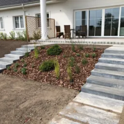 Nowoczesny ogród przy białym domu z tarasem, z betonowymi schodami prowadzącymi do rabaty wysypanej korą i obsadzonej młodymi krzewami i trawami ozdobnymi.