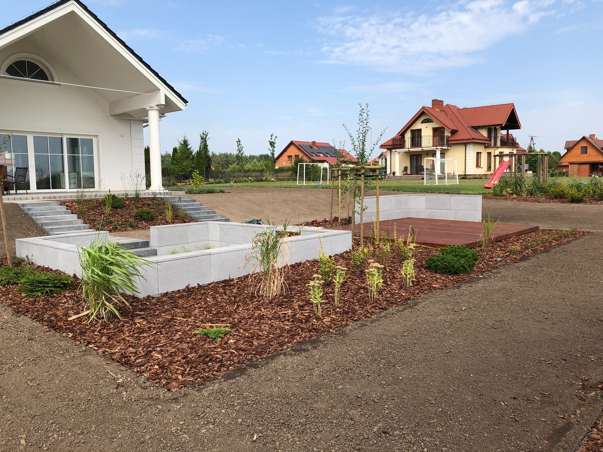 Nowoczesny ogród w trakcie realizacji: betonowe rabaty, drewniany podest, świeżo posadzone rośliny, kora rozsypana na rabatach, w tle domy jednorodzinne i plac zabaw.