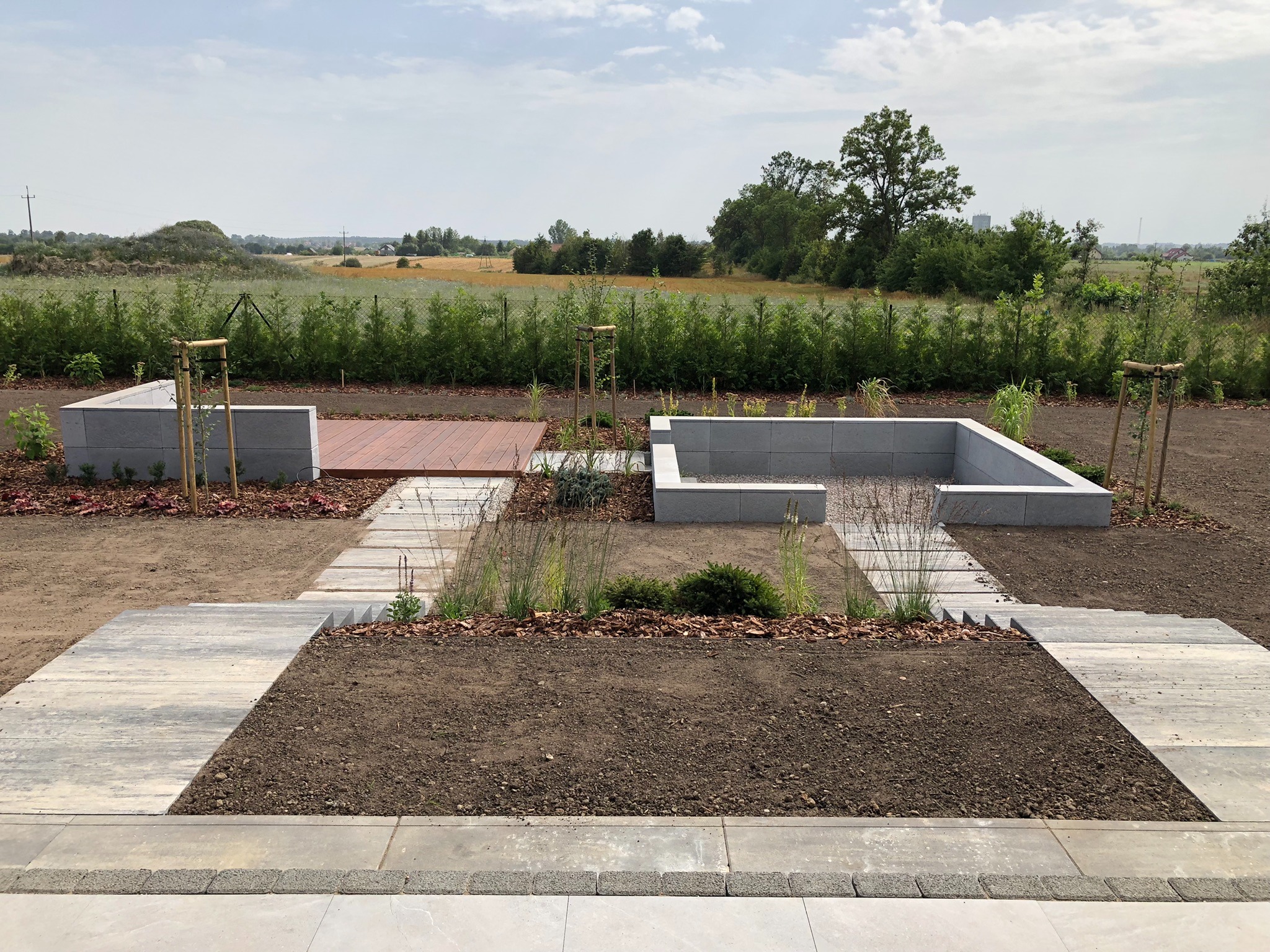 Nowoczesny ogród w trakcie realizacji, widoczne podwyższone rabaty z szarego betonu, drewniany podest, ścieżka z płyt betonowych, młode drzewka podparte bambusowymi tyczkami, przygotowana ziemia...