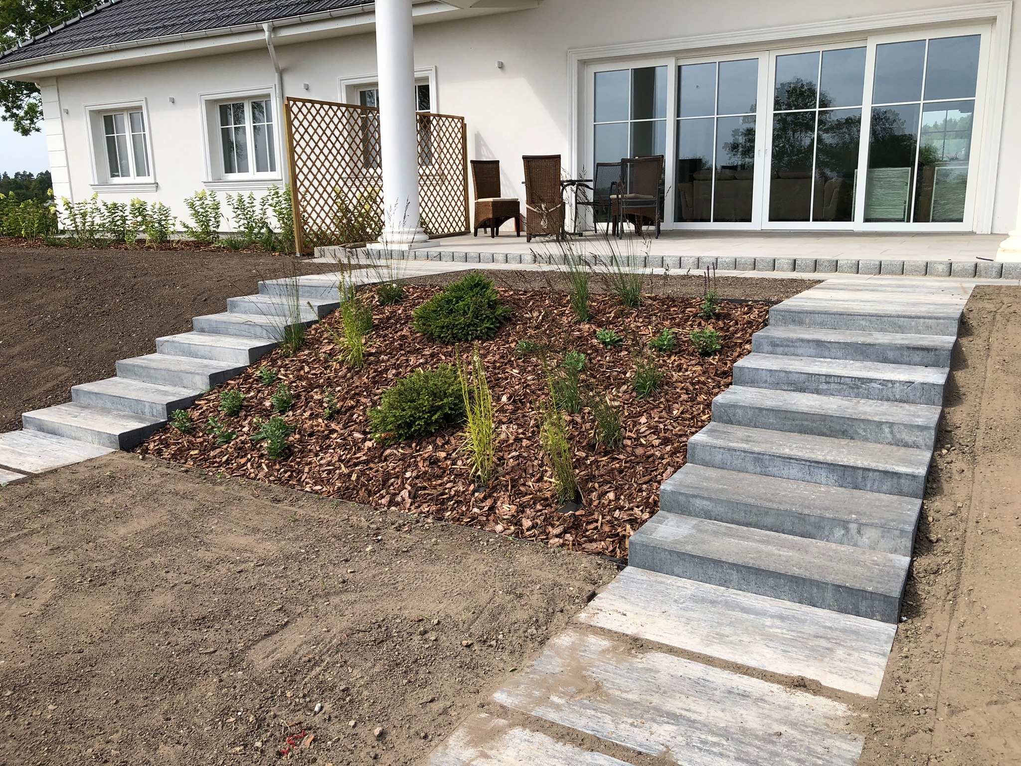 Nowoczesny ogród przy białym domu z tarasem, z betonowymi schodami prowadzącymi do rabaty wysypanej korą i obsadzonej młodymi krzewami i trawami ozdobnymi.