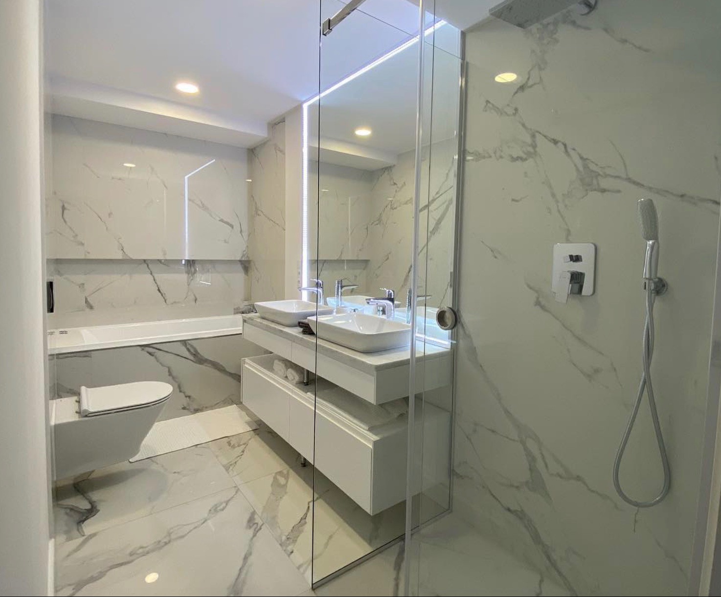 Nowoczesna łazienka z marmurowymi płytkami, podwójną umywalką nablatową na białej szafce z szufladami, kabiną prysznicową typu walk-in i wiszącą toaletą.