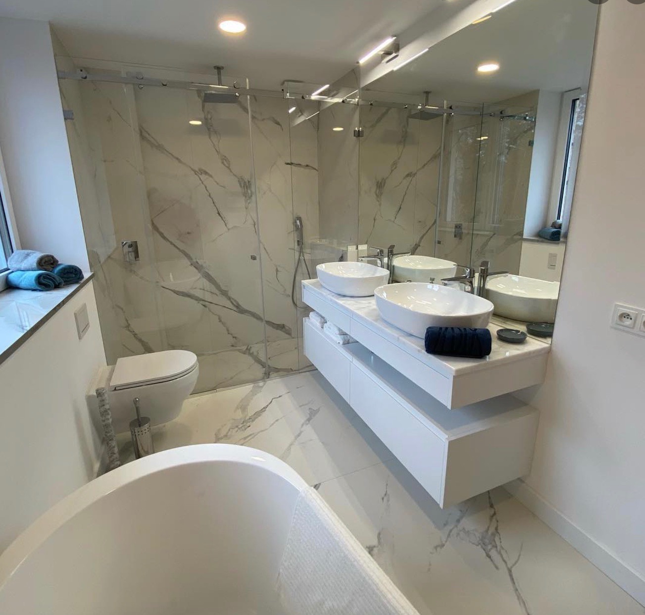 Nowoczesna, jasna łazienka z marmurowymi płytkami, przeszklonym prysznicem, podwójną umywalką na białej szafce i wolnostojącą wanną. Szare ręczniki ułożone na parapecie, czarny ręcznik na blacie.