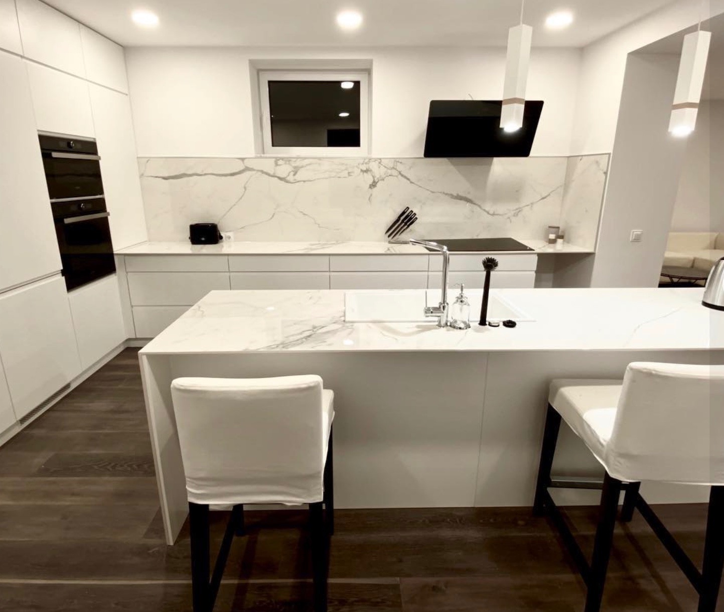 Nowoczesna, minimalistyczna kuchnia z wyspą i białymi frontami, marmurowy backsplash, czarny okap i drewniana podłoga, widoczne dwa krzesła barowe z białymi pokrowcami.