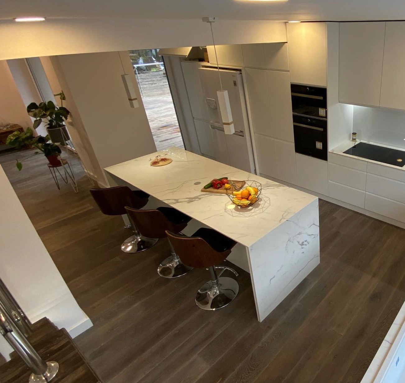 Nowoczesna kuchnia z wyspą i trzema hokerami, widok z góry, na blacie o marmurkowym wzorze leżą owoce i warzywa, podłoga z paneli imitujących drewno.