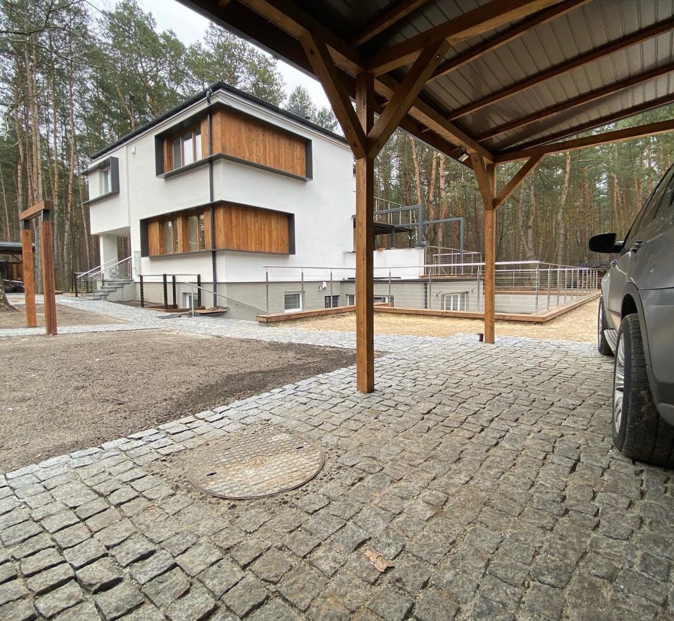 Nowoczesny dom z białymi ścianami i drewnianymi akcentami, widok z zadaszonego parkingu wyłożonego kostką brukową, w tle las.