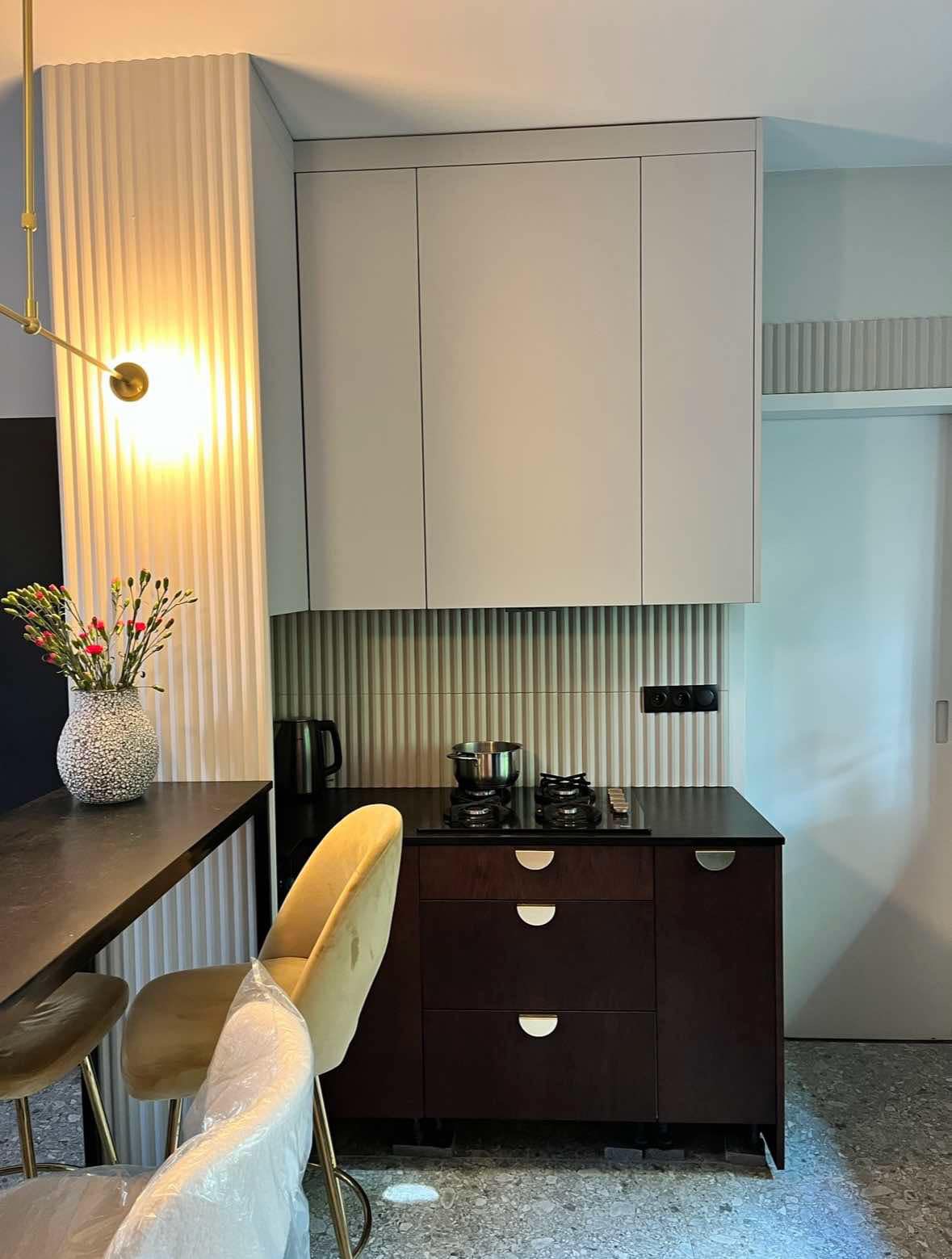 Nowoczesna kuchnia z ciemnymi szafkami dolnymi i jasnymi górnymi, ryflowana ściana, złote akcenty oświetleniowe i krzesła barowe, kwiaty w wazonie na blacie.