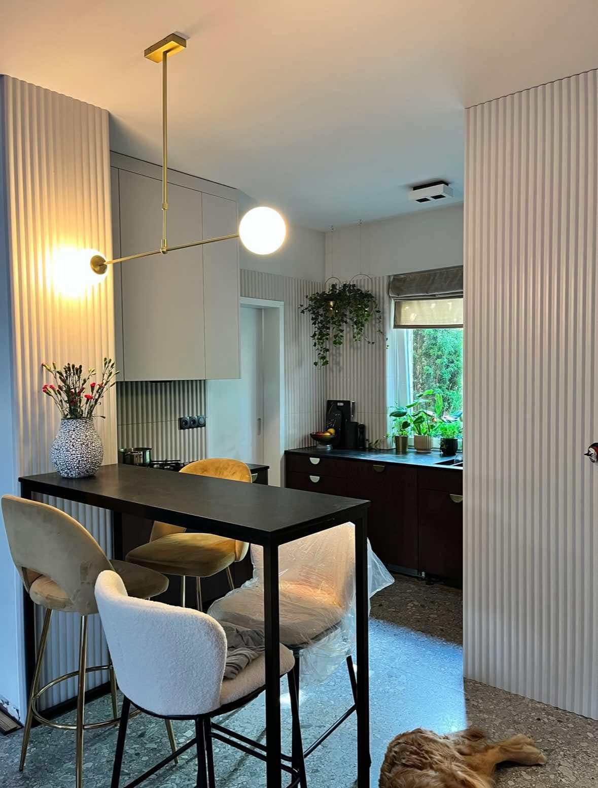 Nowoczesna kuchnia z wysokim stołem barowym, trzema krzesłami (jedno z nich w folii ochronnej), złotą lampą sufitową i ścianami z pionowymi panelami dekoracyjnymi. W tle rośliny doniczkowe...