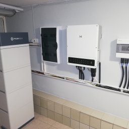 Solamis - Instalacja domowa: biały magazyn energii Pylontech, inwerter z czarnym panelem, skrzynka elektryczna i kable w korytkach na tle ściany z płytkami.