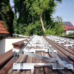 Solamis - Stelaż pod panele na brązowym dachu z blachy trapezowej. W tle zieleń drzew i budynek z panelami słonecznymi. Montaż konstrukcji wsporczej.