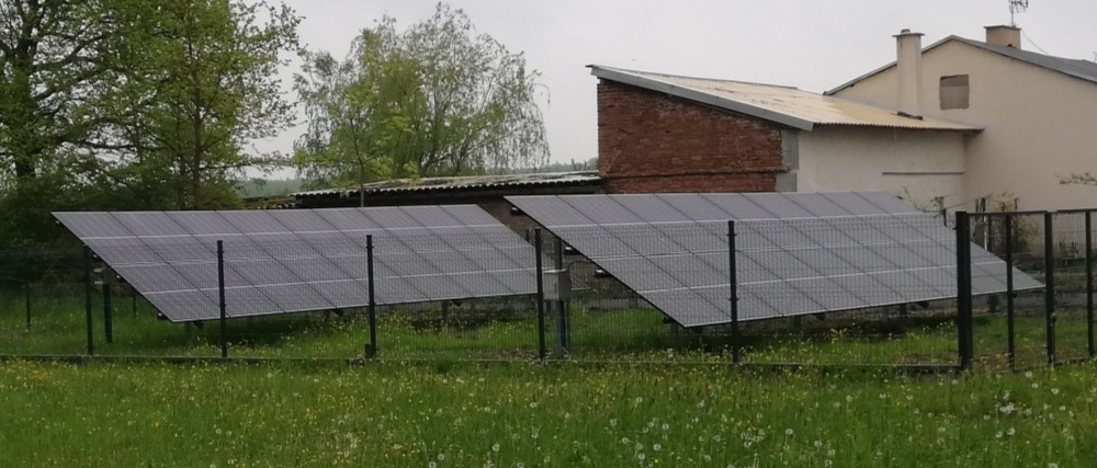 Dwa rzędy paneli fotowoltaicznych na metalowej konstrukcji, otoczone zielenią i ogrodzeniem. W tle budynki, pochmurne niebo. Widok z niskiej perspektywy.