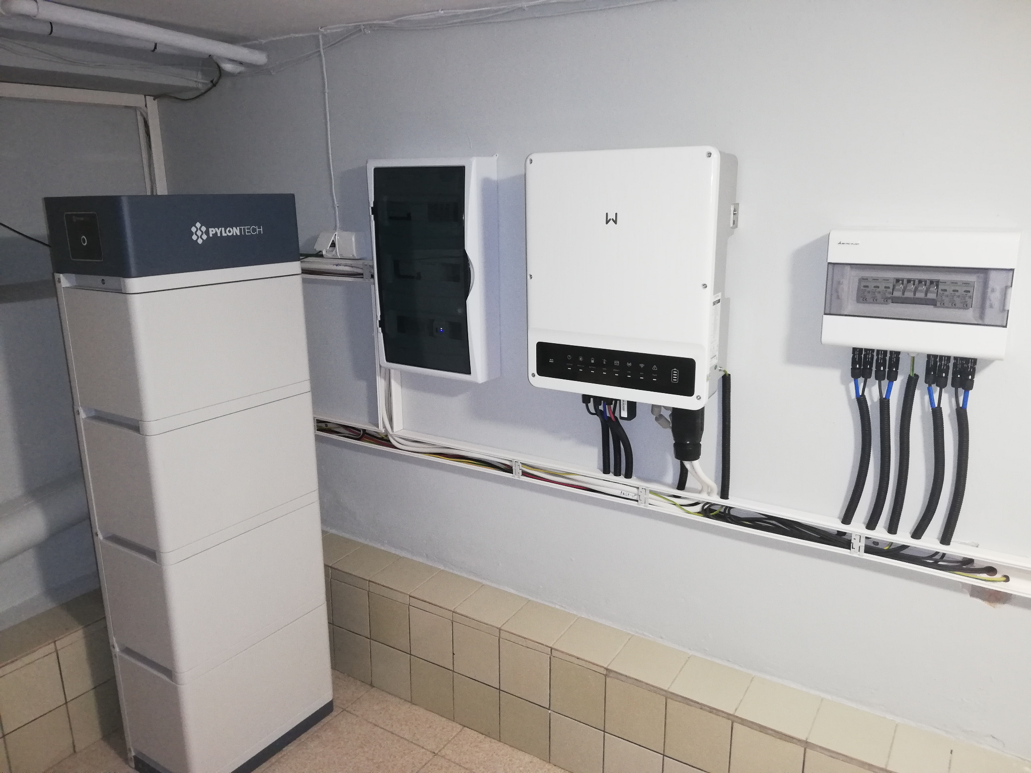 Instalacja domowa: biały magazyn energii Pylontech, inwerter z czarnym panelem, skrzynka elektryczna i kable w korytkach na tle ściany z płytkami.