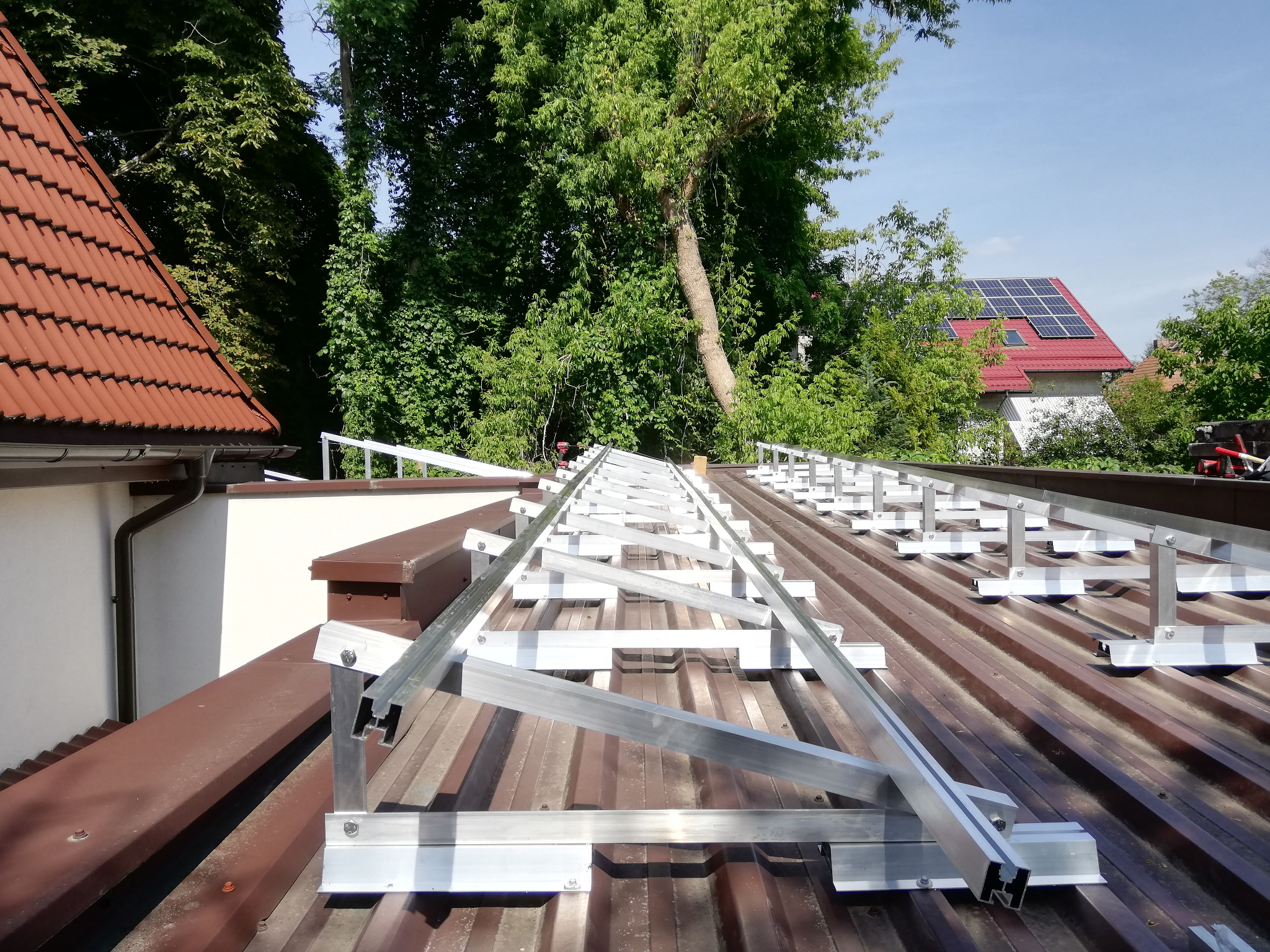 Stelaż pod panele na brązowym dachu z blachy trapezowej. W tle zieleń drzew i budynek z panelami słonecznymi. Montaż konstrukcji wsporczej.