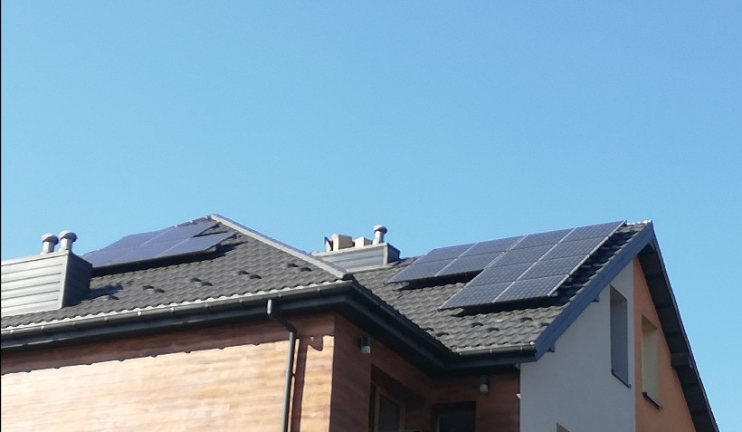 Instalacja paneli słonecznych na dachu domu jednorodzinnego w Łodzi. Widok na czarne panele fotowoltaiczne i komin na tle błękitnego nieba.