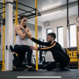 Kobieta wykonuje przysiad z jedną nogą opartą o ścianę, a mężczyzna w czarnej bluzie z pomarańczowymi paskami kontroluje poprawność ruchu w siłowni.