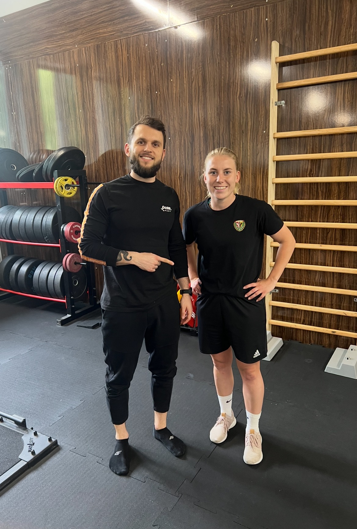 Uśmiechnięty mężczyzna z tatuażem na przedramieniu, w czarnej koszulce sportowej, wskazuje na równie uśmiechniętą kobietę w czarnym sportowym stroju stojącą obok, w tle drabinki gimnastyczne...