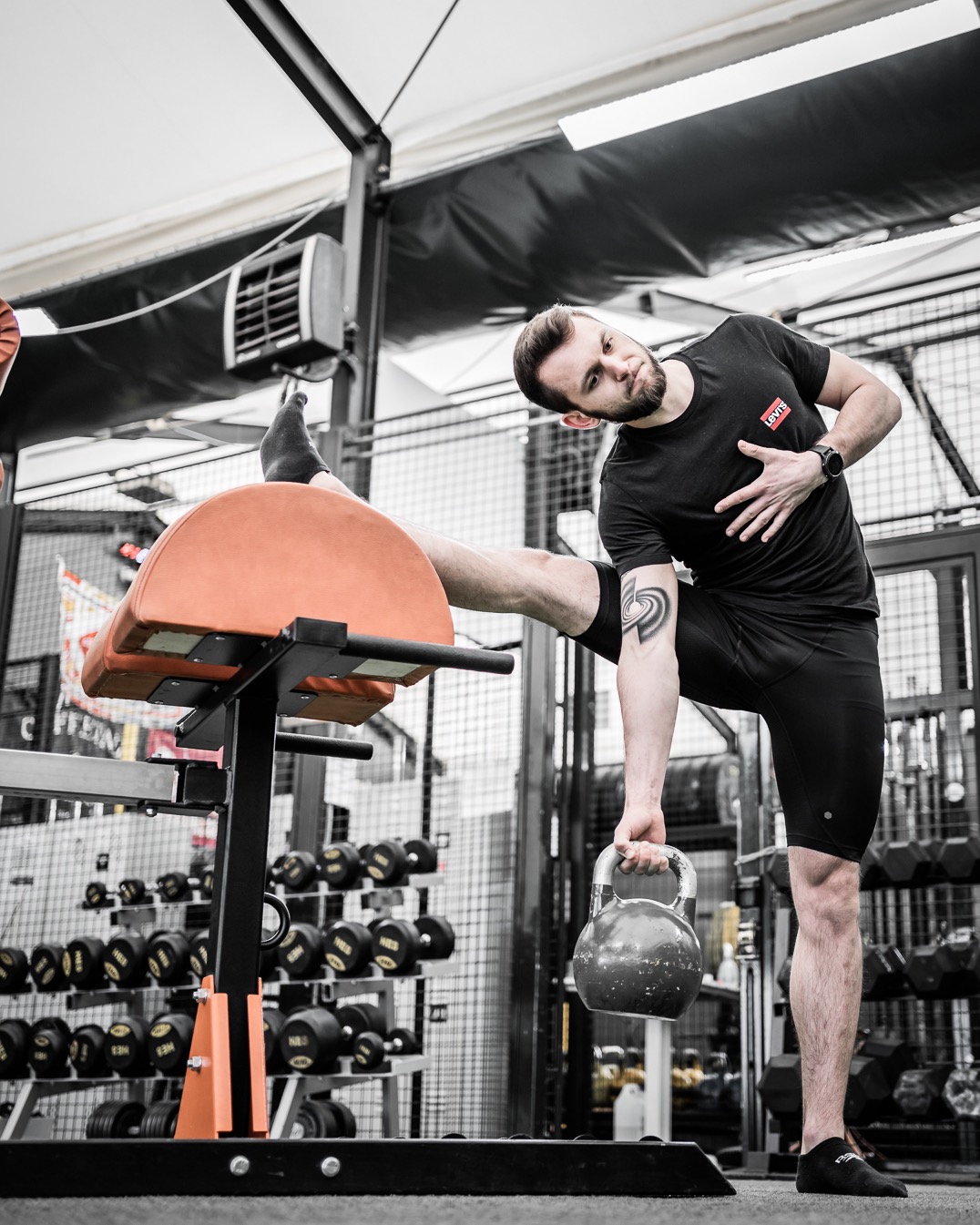 Mężczyzna w czarnym stroju sportowym rozciąga nogę na pomarańczowym sprzęcie do ćwiczeń, trzymając kettlebell w siłowni z rzędem hantli w tle.