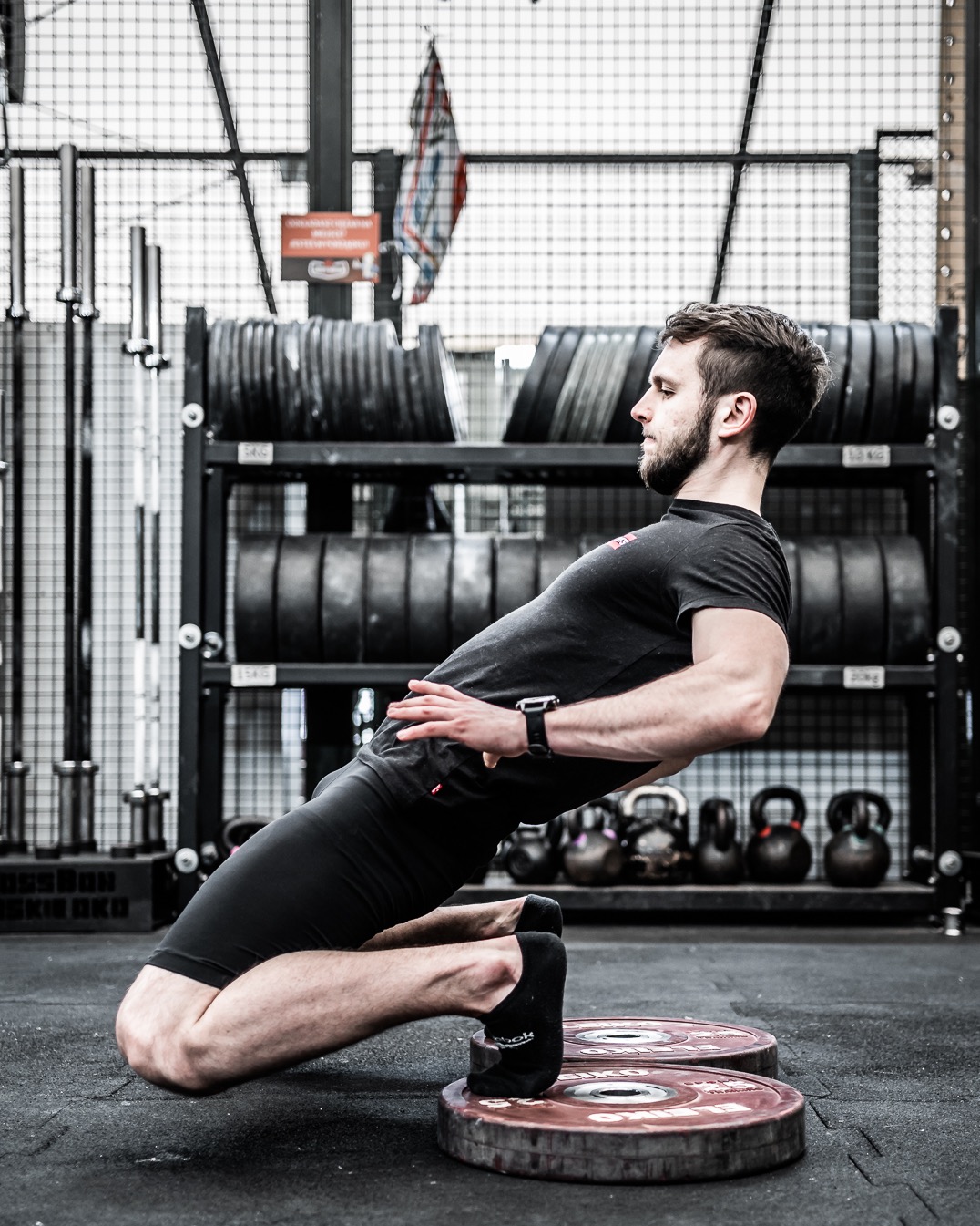 Mężczyzna w czarnym stroju wykonuje ćwiczenie kolan na dwóch obciążeniach w siłowni, z widocznym stojakiem na ciężary i kettlebells w tle.