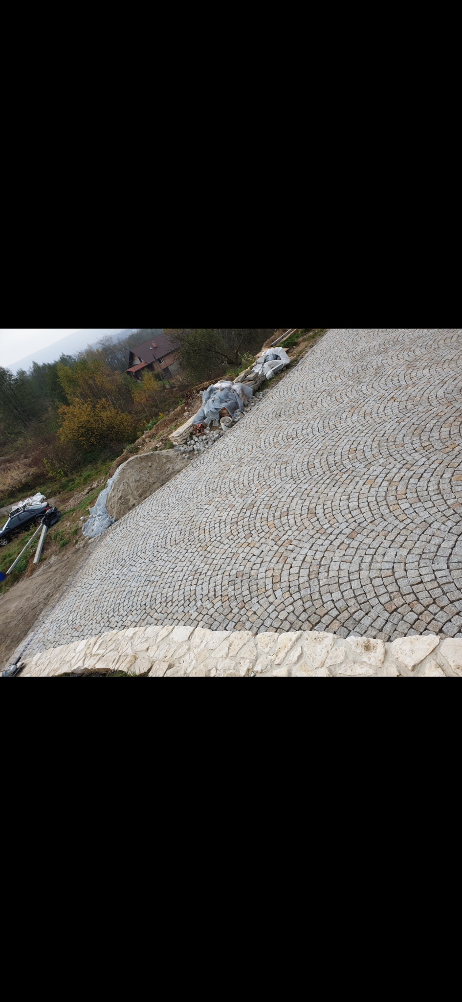 Ułożona kostka granitowa w okrągły wzór na pochyłym terenie, z widocznymi przygotowanymi materiałami i częściowo widocznym domem w tle.
