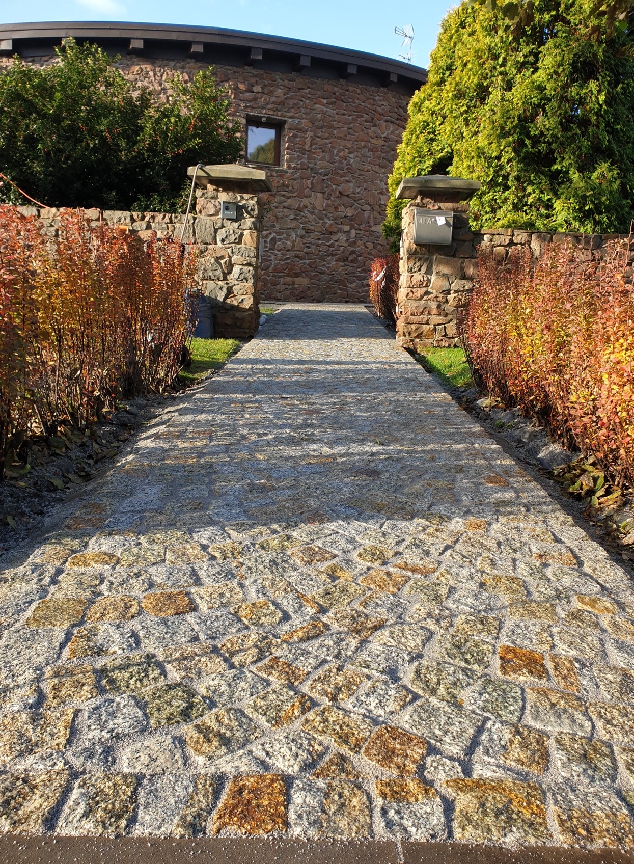 Ścieżka z nieregularnej kostki w odcieniach szarości i brązu prowadząca do kamiennego domu, otoczona rdzawymi krzewami i kamiennymi słupkami.